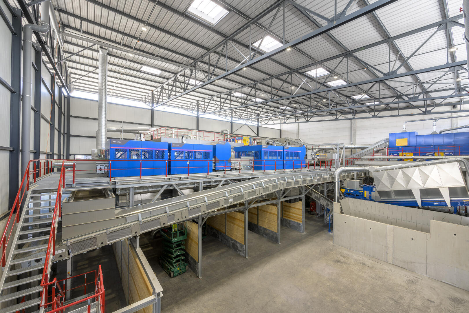 Eine geräumige Industrieanlage mit Metallstegen, roten Geländern, blauen Maschinen und großen Förderbändern zum Sortieren oder Verarbeiten von Materialien, alles unter einer hohen, gut beleuchteten Decke.