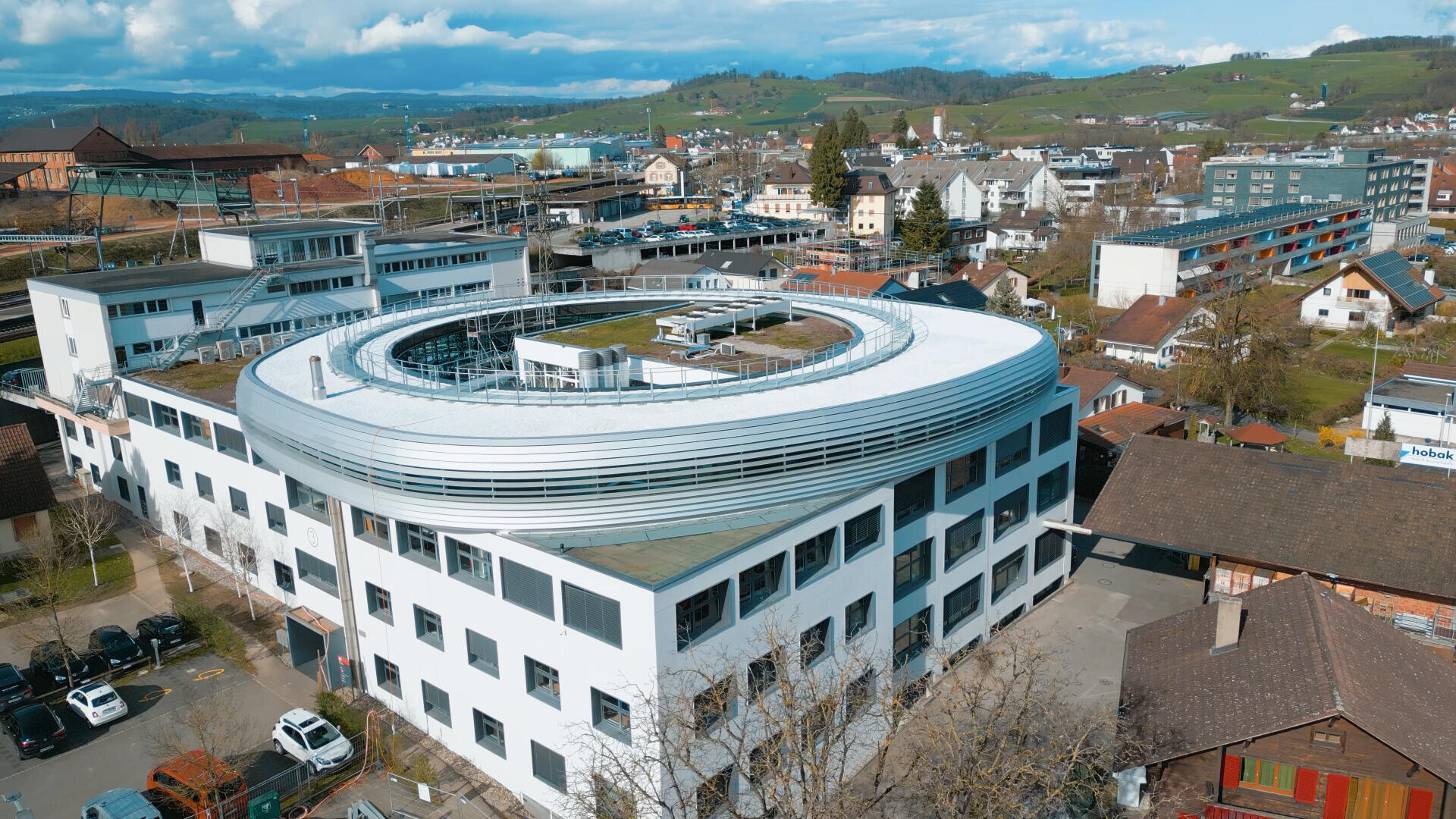 Luftaufnahme eines modernen weißen Gebäudes mit kreisförmigem Dach und grünem Dach, umgeben von kleineren Gebäuden, Bäumen und einer hügeligen Landschaft im Hintergrund.