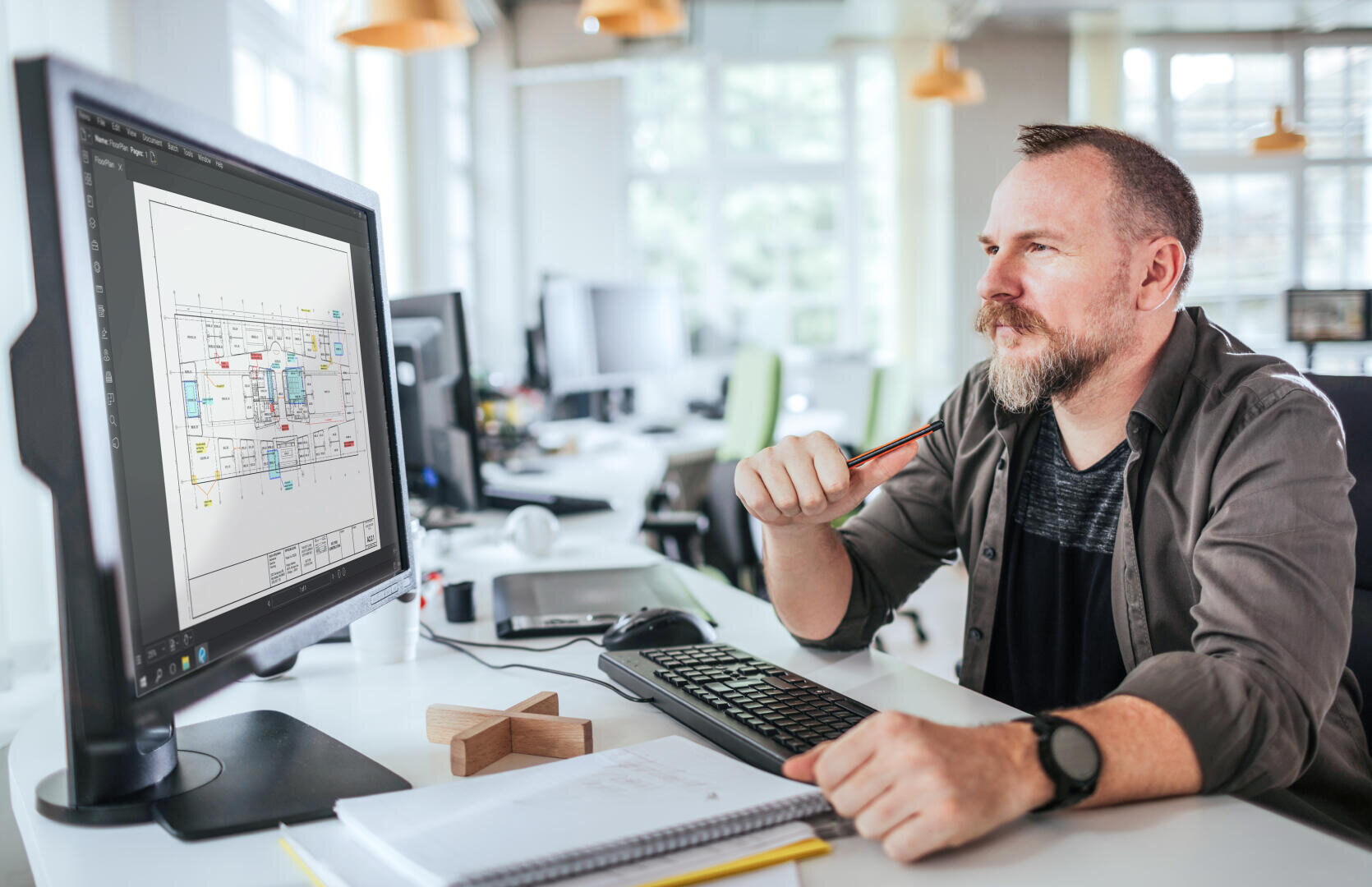 Ein Mann mit Bart sitzt an einem Schreibtisch in einem modernen Büro und betrachtet nachdenklich die auf einem Computermonitor angezeigten Architekturpläne. Auf dem Schreibtisch liegen ein Notizbuch, ein Bleistift und andere Büroutensilien.