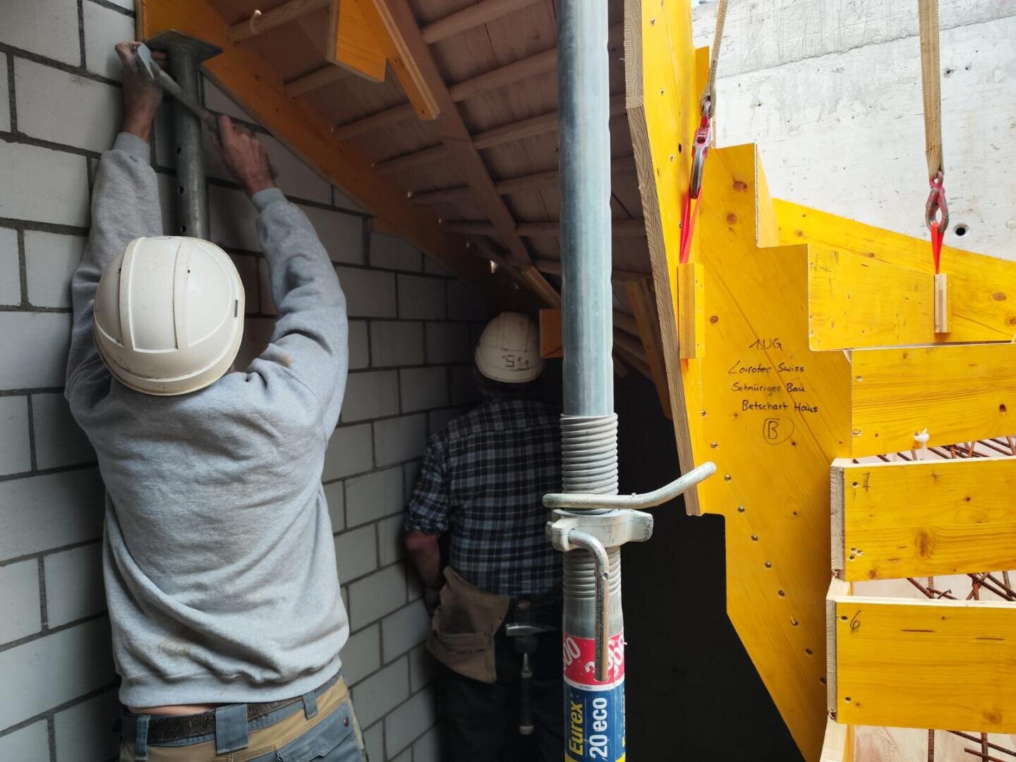 Zwei Bauarbeiter mit Helmen arbeiten unter einem Schrägdach; einer richtet eine Metallstange neben einer Wand aus, während der andere den Blick abwendet. Rechts ist eine leuchtend gelbe Holzschalung zu sehen.