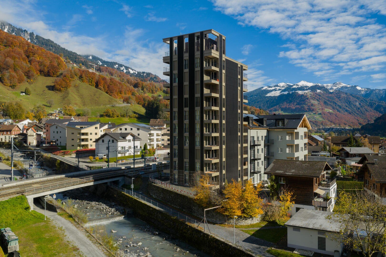 Ein hohes modernes Gebäude steht in einer kleinen Stadt, umgeben von traditionellen Häusern, grünen Hügeln und schneebedeckten Bergen unter einem teilweise bewölkten Himmel. Neben den Gebäuden verlaufen ein Fluss und eine Brücke.