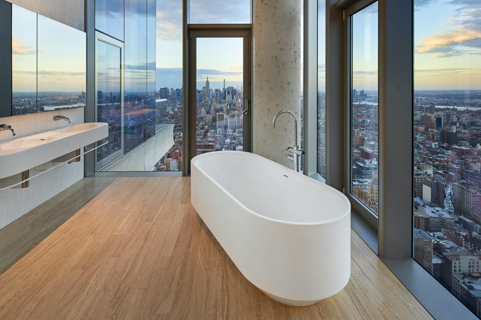 Modernes Badezimmer mit einer freistehenden ovalen Badewanne und einem Doppelwaschbecken. Die raumhohen Fenster bieten einen Panoramablick auf die Skyline der Stadt bei Sonnenuntergang. Helle Holzfußböden und ein klares, minimalistisches Design schaffen ein luxuriöses Ambiente.