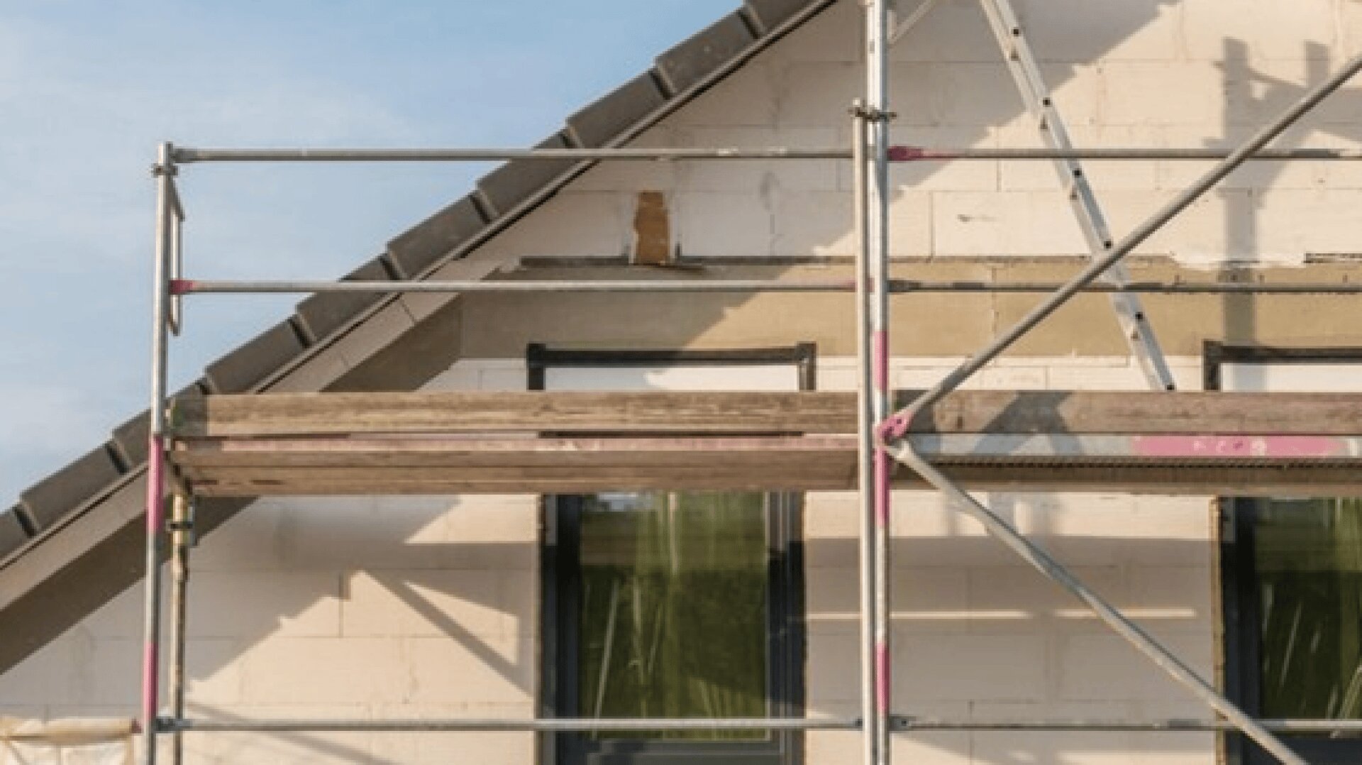 Vor einem im Bau oder in der Renovierung befindlichen Haus ist ein Baugerüst aufgebaut. Das Äußere des Hauses ist unfertig, und es sind zwei Fenster zu sehen, die teilweise durch das Gerüst verdeckt sind.