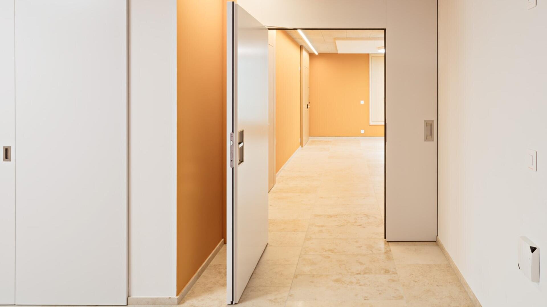 Ein moderner, minimalistischer Flur mit beigem Fliesenboden, orangefarbenen Akzentwänden und weißen Schiebetüren, von denen eine teilweise geöffnet ist und einen gut beleuchteten Raum dahinter freigibt.