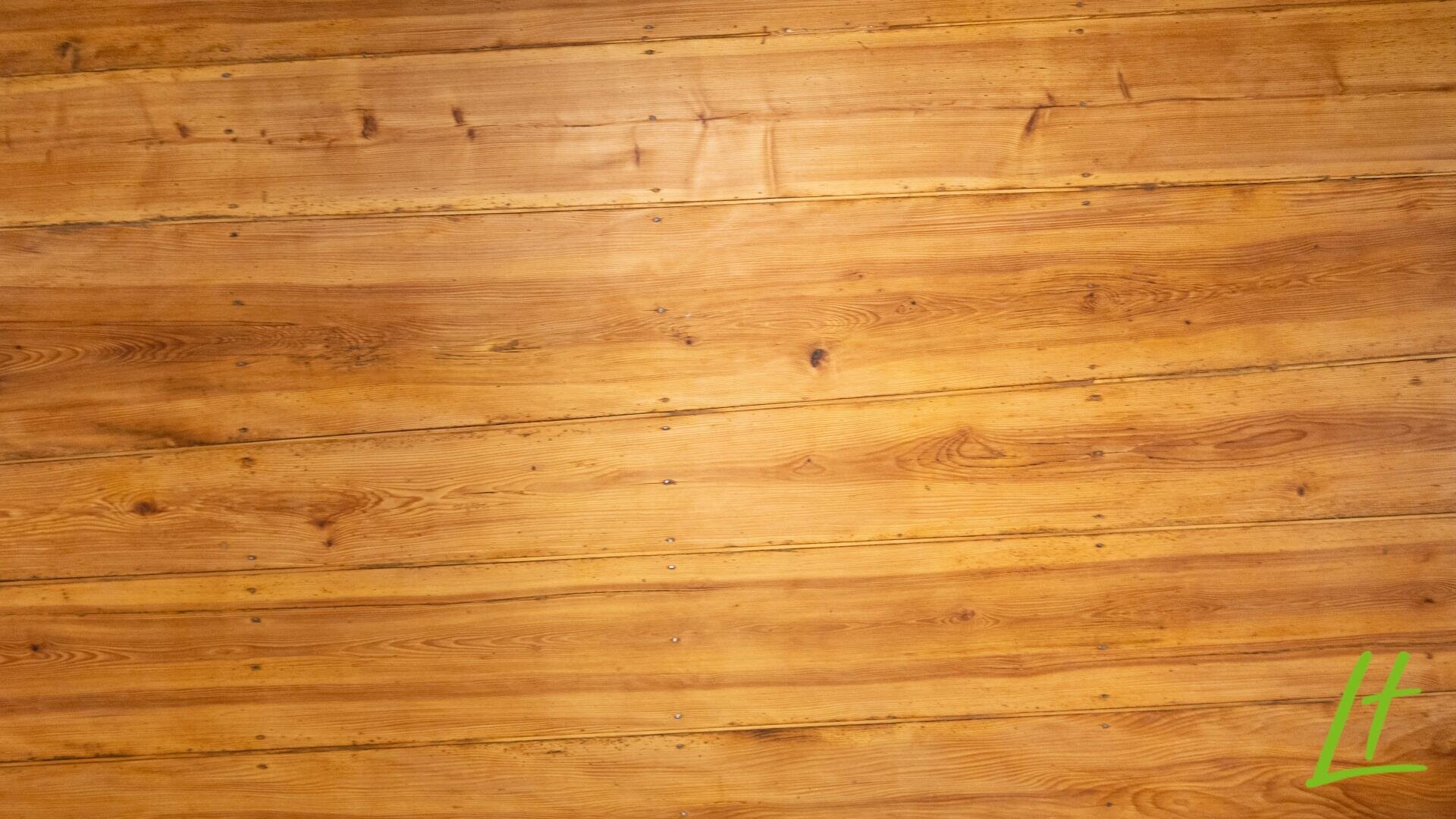 Holzplatten mit einer warmen, natürlichen Oberfläche und sichtbarer Maserung verlaufen horizontal über das Bild. In der unteren rechten Ecke befindet sich ein kleines, grünes, stilisiertes "L".