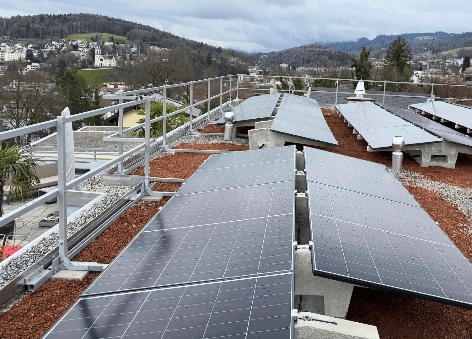 Reihen von Solarmodulen, die auf einem Dach mit Sicherheitsgeländer installiert sind, mit Blick auf eine hügelige Landschaft und eine entfernte Stadt unter einem bewölkten Himmel.