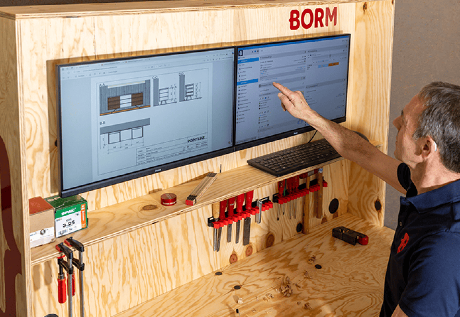 Ein Mann zeigt auf einen an der Wand montierten Computermonitor, auf dem eine Projektmanagement-Software angezeigt wird, daneben ein weiterer Monitor, auf dem Architekturzeichnungen zu sehen sind, und darunter ein hölzerner Arbeitsplatz mit Werkzeugen und Klemmen.
