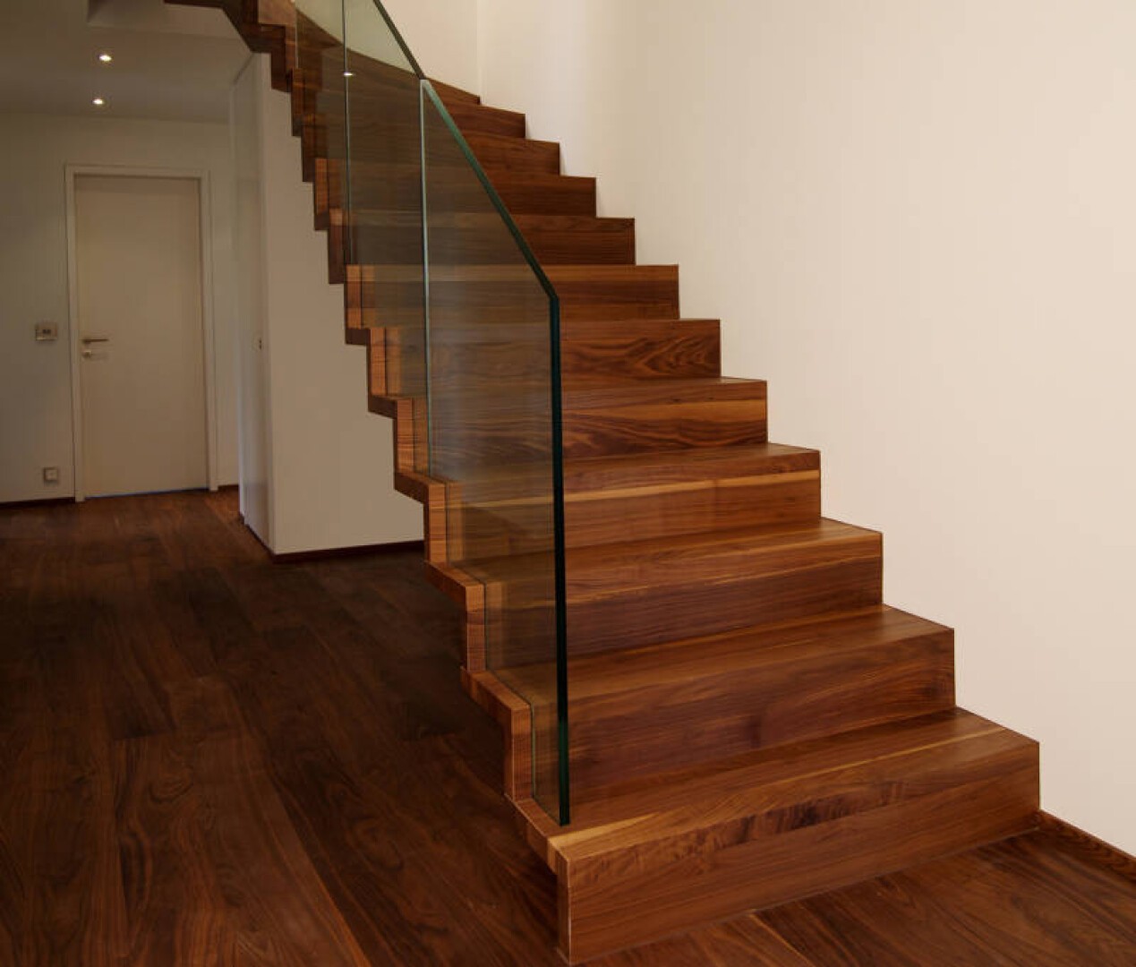 Eine moderne Treppe aus dunklem Holz mit Glasgeländer führt von einem Flur mit passendem Holzboden und schlichten weißen Wänden nach oben. Am Ende des Flurs ist eine geschlossene weiße Tür zu sehen.