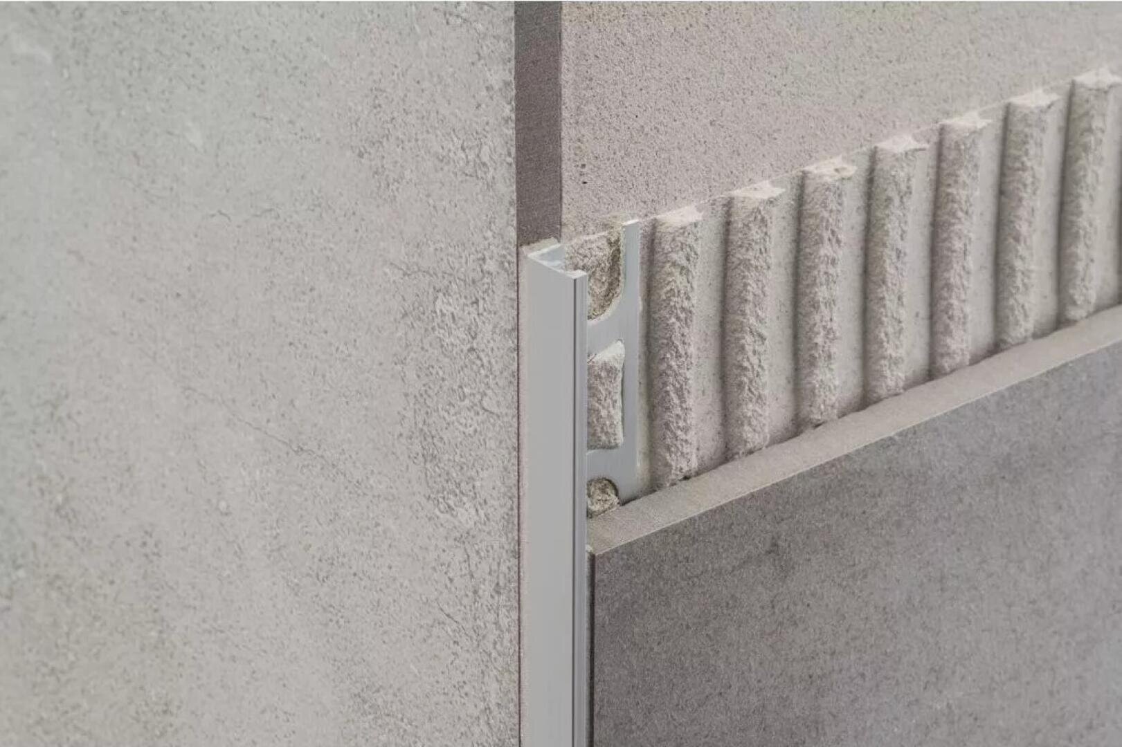 Nahaufnahme von zwei Beton- oder Steinfliesen, die an einer Ecke aufeinandertreffen, mit einer Metallleiste zwischen ihnen und einem geriffelten Kleber, der hinter der rechten Fliese sichtbar ist.
