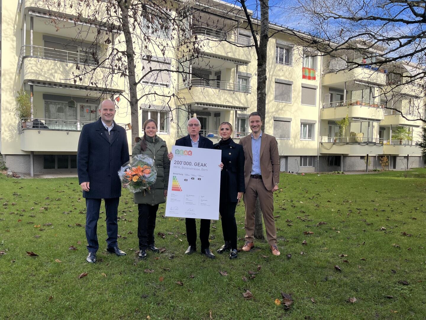 Cinq personnes se tiennent sur une pelouse devant une maison d'habitation. Trois d'entre elles tiennent un grand certificat, une personne un bouquet de fleurs. En arrière-plan, on voit des arbres sans feuilles.