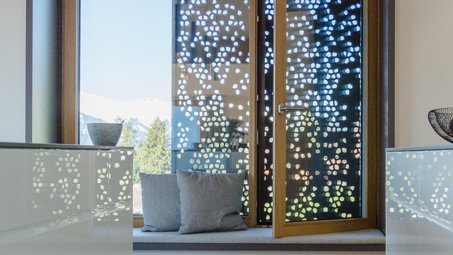 Une fenêtre moderne dotée d'un pare-vue perforé laisse entrer la lumière à motifs. Des coussins et un bol décoratif sont posés sur le rebord de la fenêtre, tandis que des pins et des montagnes enneigées sont visibles à l'extérieur.