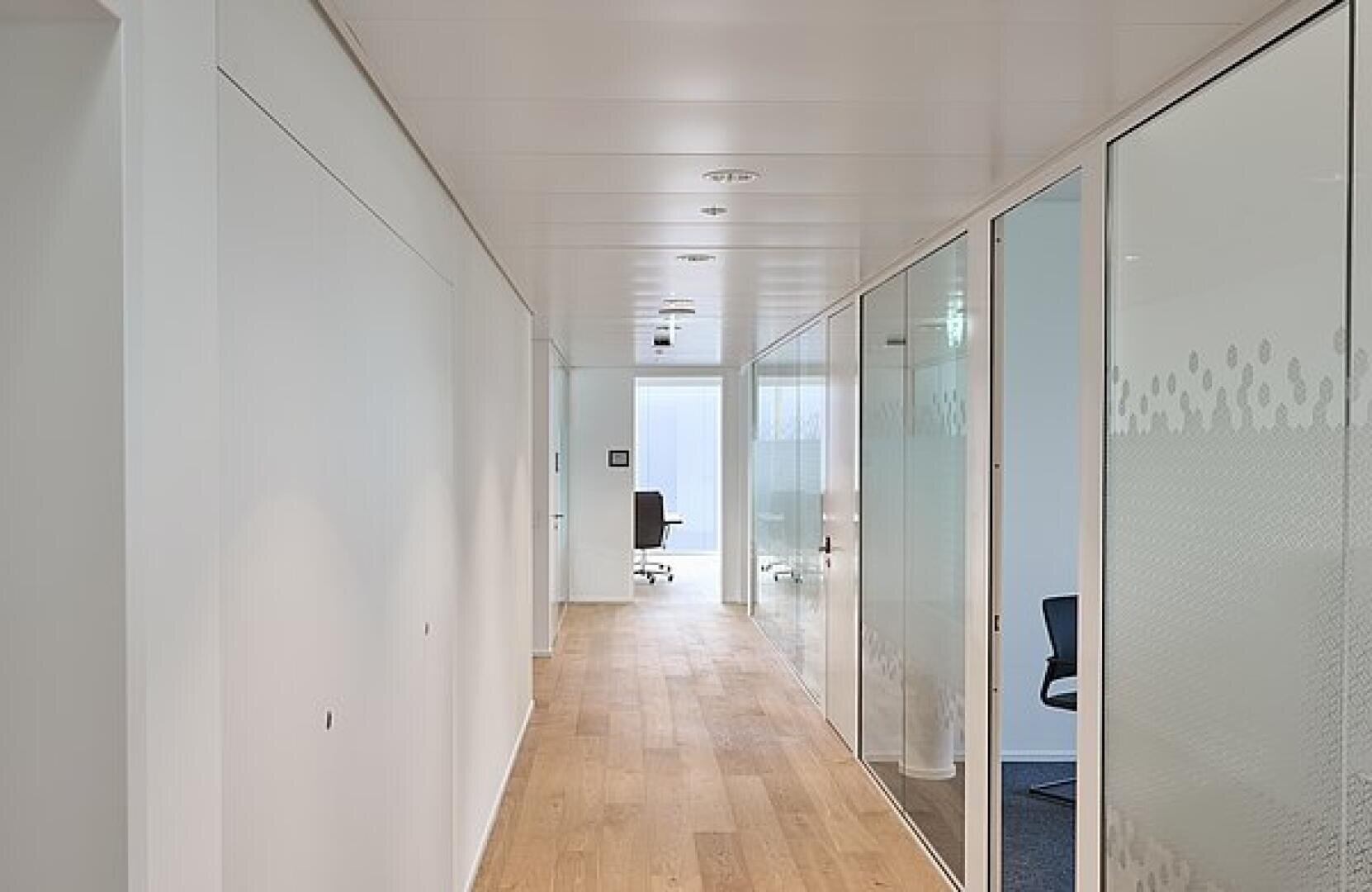 Ein moderner Büroflur mit Holzfußboden, weißen Wänden und Glastrennwänden. Einige Büros sind durch das Glas hindurch sichtbar und mit Stühlen und Schreibtischen ausgestattet. Durch ein Fenster am Ende des Flurs fällt natürliches Licht in den Flur.