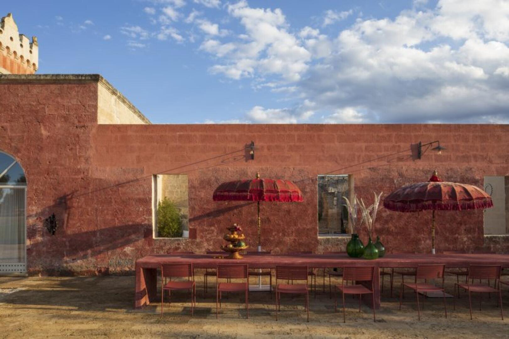 Une longue table de salle à manger d'extérieur avec des chaises rouges est assise contre un mur rustique en terre cuite. Deux parapluies rouges ornés font de l'ombre, et des vases décoratifs ainsi qu'un plateau à étages décorent la table sous un ciel partiellement nuageux.
