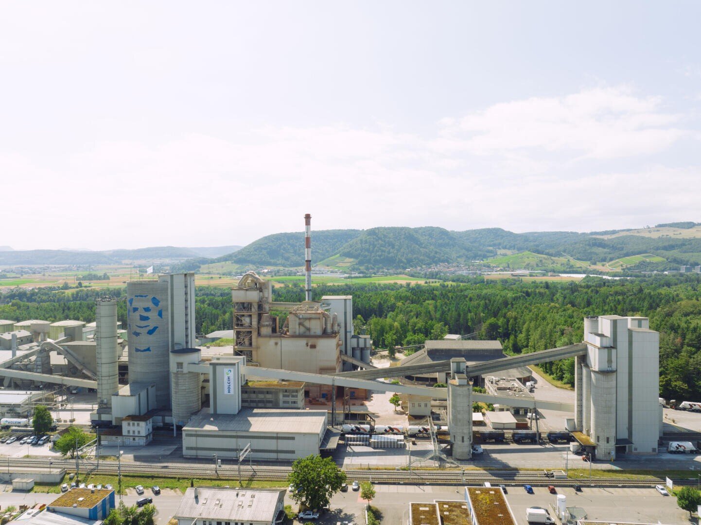 Eine große industrielle Zementfabrik mit hohen Silos und einem zentralen Schornstein, umgeben von grünen Bäumen und Hügeln unter einem hellen, teilweise bewölkten Himmel. Förderbänder verbinden mehrere Gebäude des Komplexes miteinander.