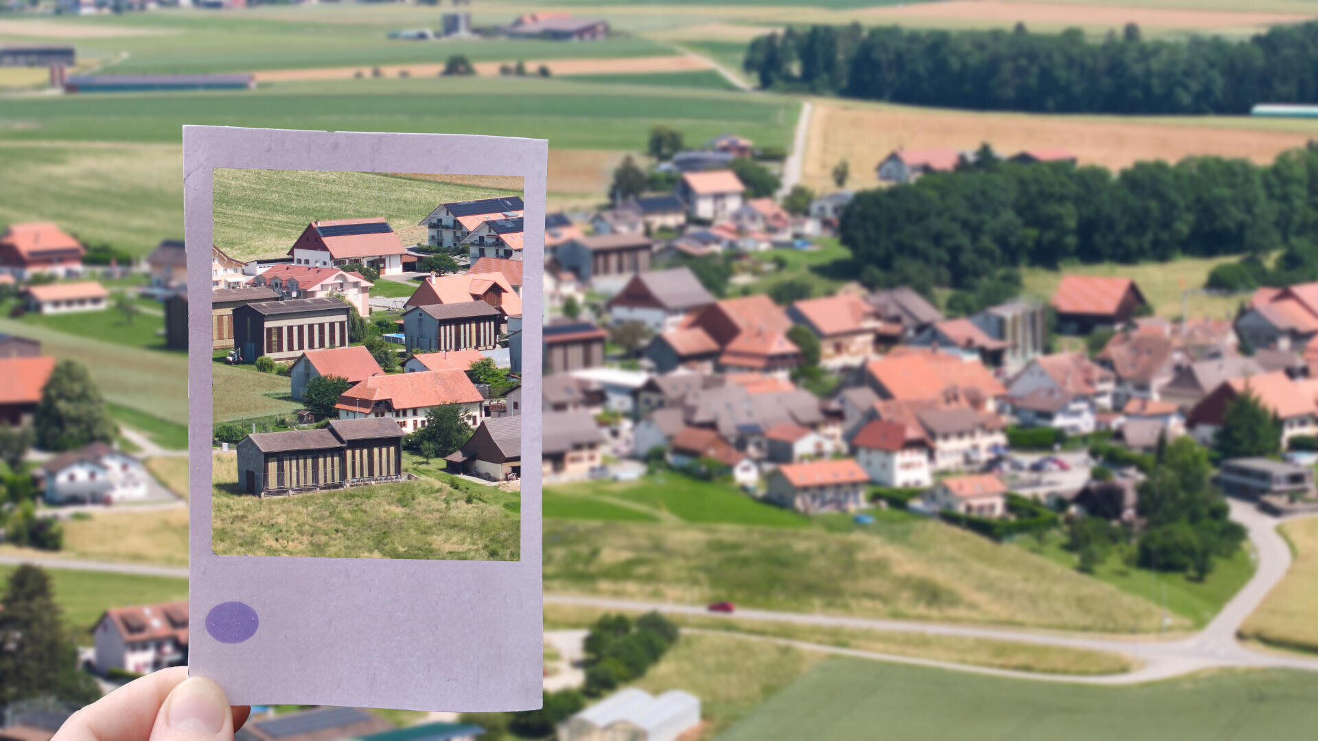 Eine Hand hält ein Foto, das einen Teil eines ländlichen Dorfes zeigt, das mit denselben Gebäuden und der Landschaft im unscharfen Hintergrund der tatsächlichen Dorfszene übereinstimmt. Sowohl auf dem Foto als auch im Hintergrund sind Felder und Häuser zu sehen.