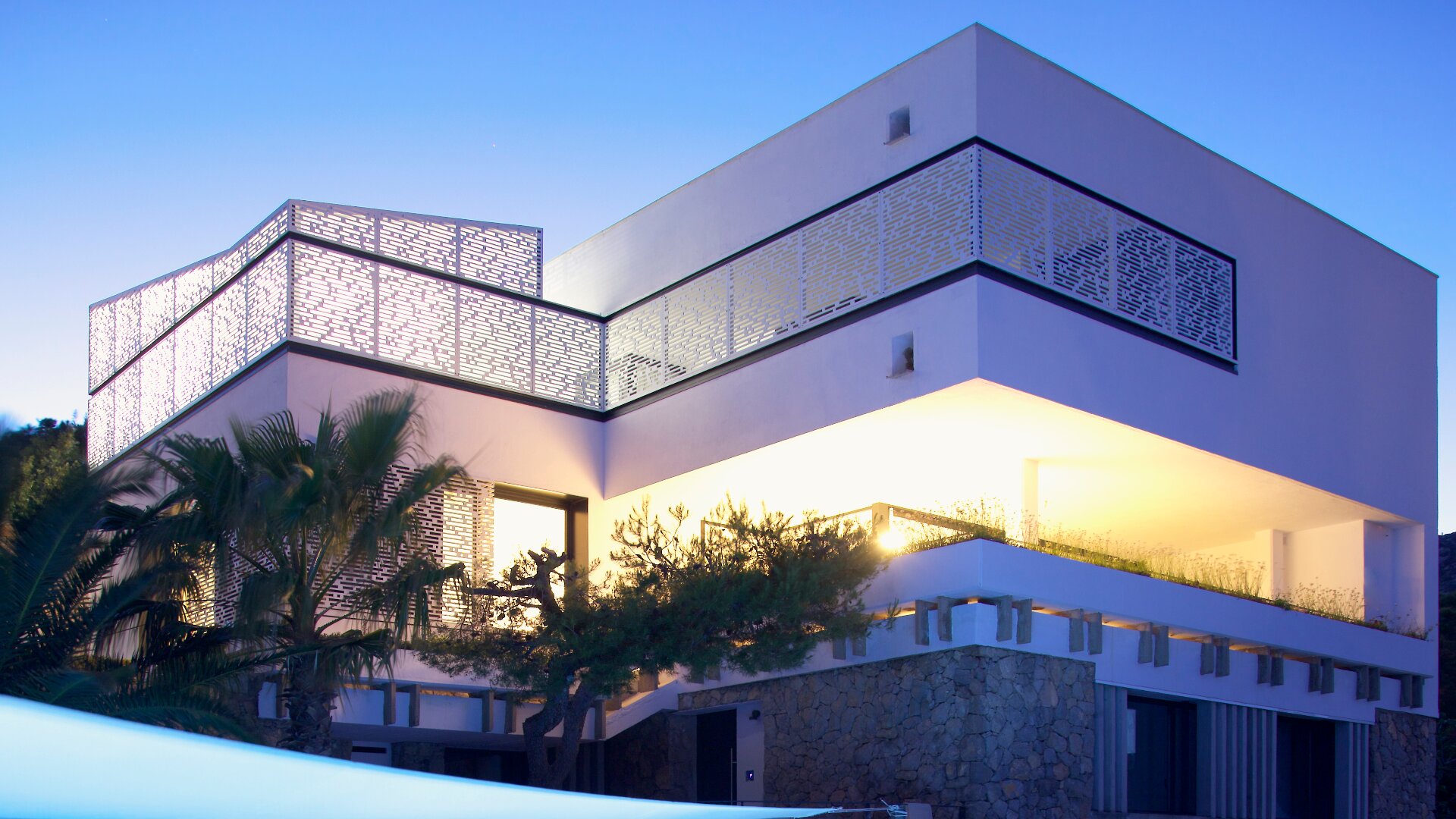 Modernes zweistöckiges Haus in der Abenddämmerung mit beleuchteten geometrischen Metallgeländern, großen Fenstern, Steinakzenten und umgeben von Bäumen und Pflanzen. Der Himmel ist klar und blau.