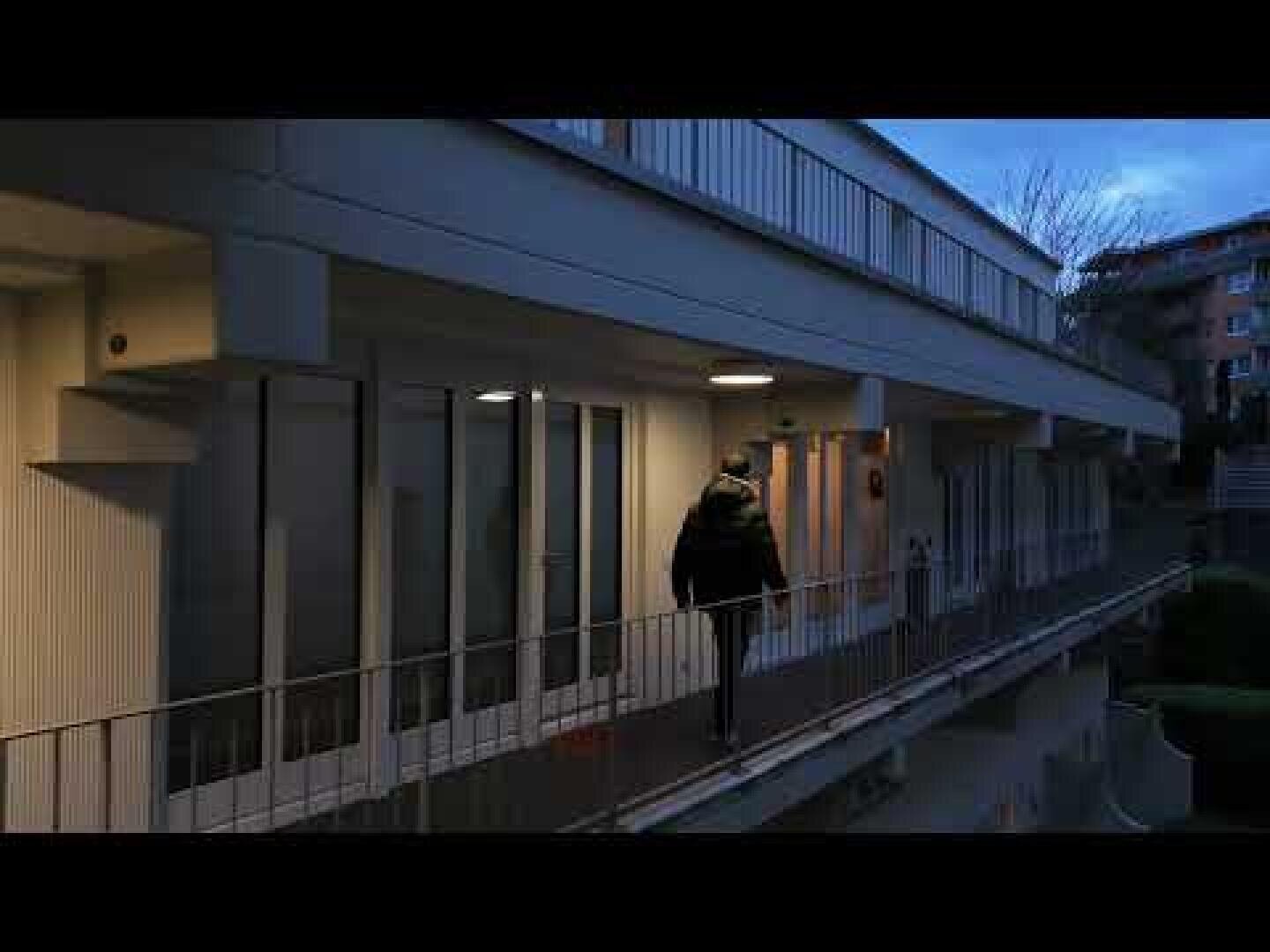 Eine dunkel gekleidete Person geht in der Abenddämmerung allein über einen Außengang eines modernen Gebäudes, der von warmen Lichtern beleuchtet wird.