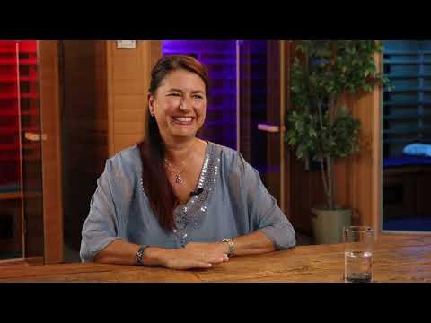 Une femme aux longs cheveux bruns, vêtue d'un chemisier bleu clair, est assise à une table en bois à l'intérieur, en souriant. Un verre d'eau est posé sur la table, et des lumières colorées et floues ainsi qu'une plante en pot se trouvent à l'arrière-plan.