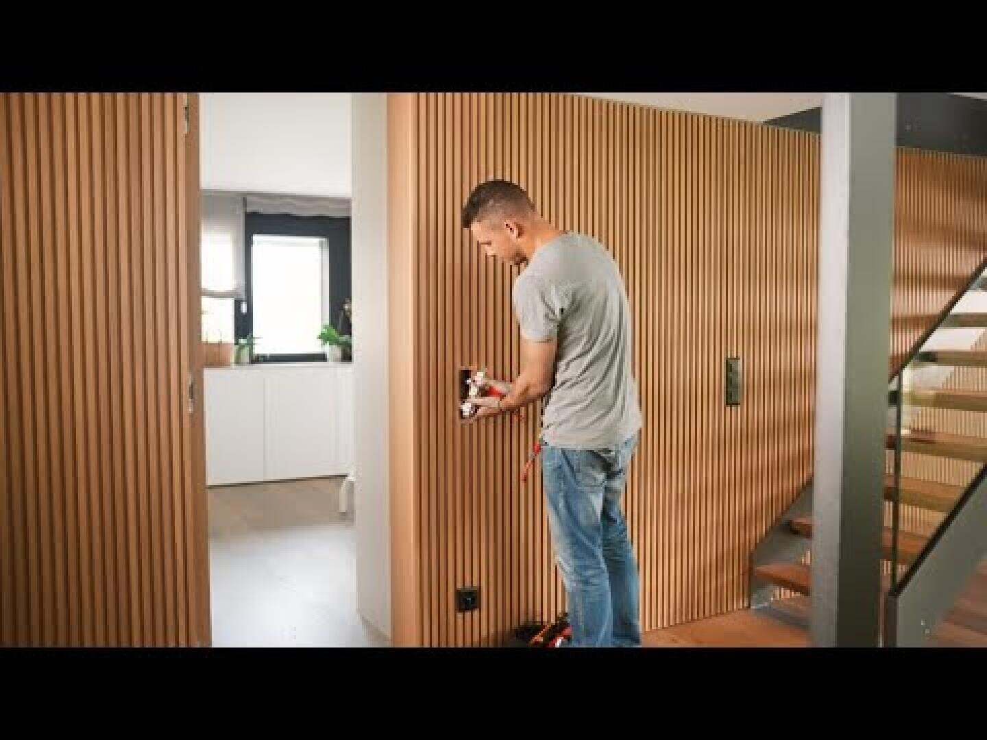 Eine Person steht neben einer Holzlattenwand in einem modernen Haus, hält Elektrowerkzeuge in der Hand und arbeitet in der Nähe einer Steckdose. Im Hintergrund sind eine Küche und ein Treppenhaus zu sehen.