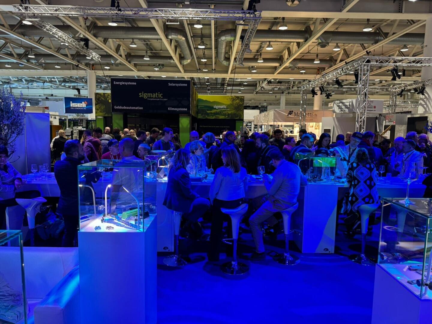 Eine große Gruppe von Menschen sitzt und steht um Stehtische auf einer belebten Messe oder Ausstellung, umgeben von beleuchteten Vitrinen und verschiedenen Firmenständen in einer geräumigen, gut beleuchteten Halle.