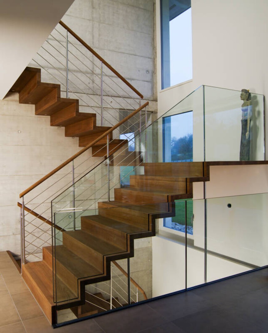 Escalier moderne en bois avec garde-corps en verre et métal dans une maison contemporaine, avec de grandes fenêtres et une sculpture sur le palier. Le béton et les murs blancs complètent le design minimaliste.