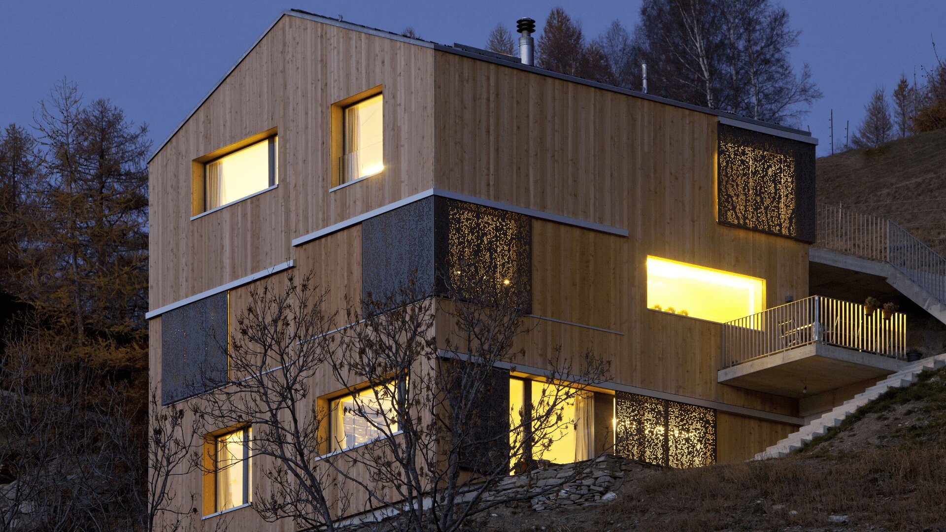 Ein modernes dreistöckiges Holzhaus mit großen Fenstern, die in der Abenddämmerung leuchten, umgeben von Bäumen und auf einem abfallenden Hügel gelegen, mit einer Metalltreppe und dekorativen Metallpaneelen an der Außenseite.
