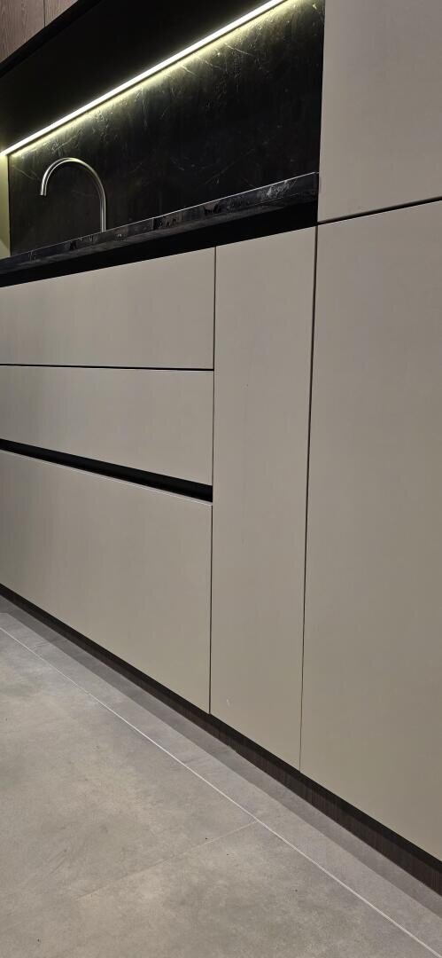 Cuisine moderne avec des armoires beige mat, un comptoir foncé, un éclairage sous l'armoire, un évier encastré et un sol carrelé gris clair. Le design est minimaliste et épuré.