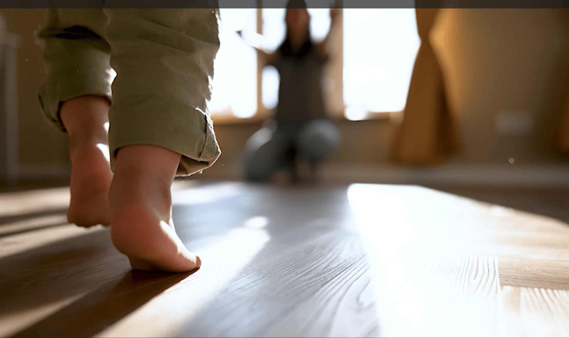 Nahaufnahme eines Kindes, das mit nackten Füßen auf einem sonnenbeschienenen Holzboden läuft, mit einem Erwachsenen im Hintergrund, der die Arme zur Ermutigung ausbreitet, unscharf.