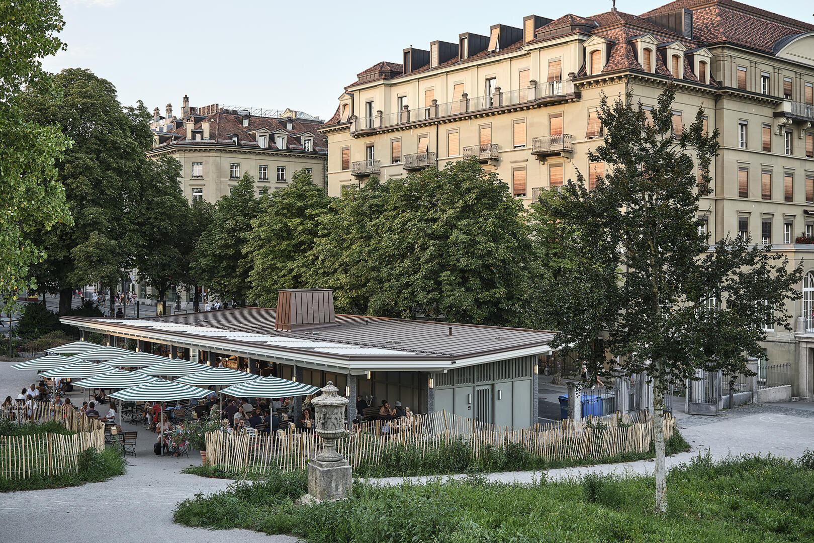 Ein modernes Straßencafé mit gestreiften Sonnenschirmen sitzt zwischen Bäumen vor einem großen historischen Gebäude mit vielen Fenstern und einem roten Ziegeldach. Es wird gegessen, und ein niedriger Holzzaun umgibt das Café.