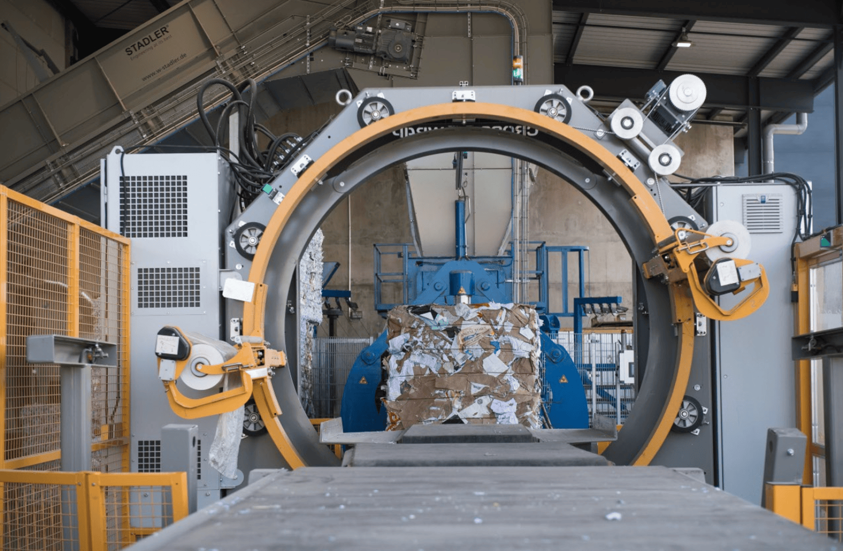 Eine große Industriemaschine komprimiert einen Stapel Karton und Papier für das Recycling in einer Fabrikumgebung, umgeben von Metallstrukturen, Förderbändern und Schutzzäunen.