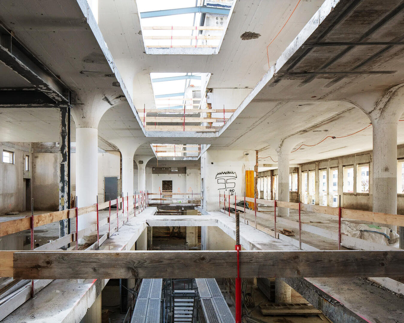 Ein leeres, teilweise abgerissenes Gebäudeinneres mit Sichtbeton, Pfeilern, offenen Decken und Bauabsperrungen. Sonnenlicht strömt durch große rechteckige Oberlichter darüber. An einer entfernten Wand ist ein Graffiti zu sehen.