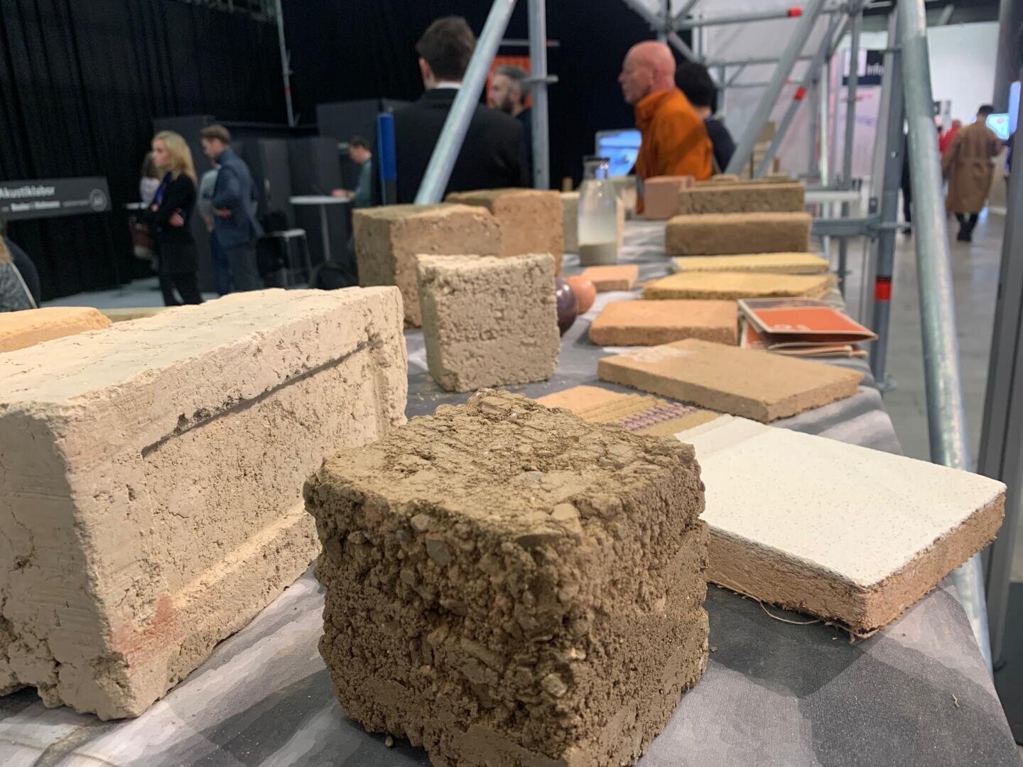 Ein Tisch mit verschiedenen Ziegelsteinen und Baustoffmustern, im Hintergrund stehen Menschen und unterhalten sich auf einer Indoor-Ausstellung oder Veranstaltung.