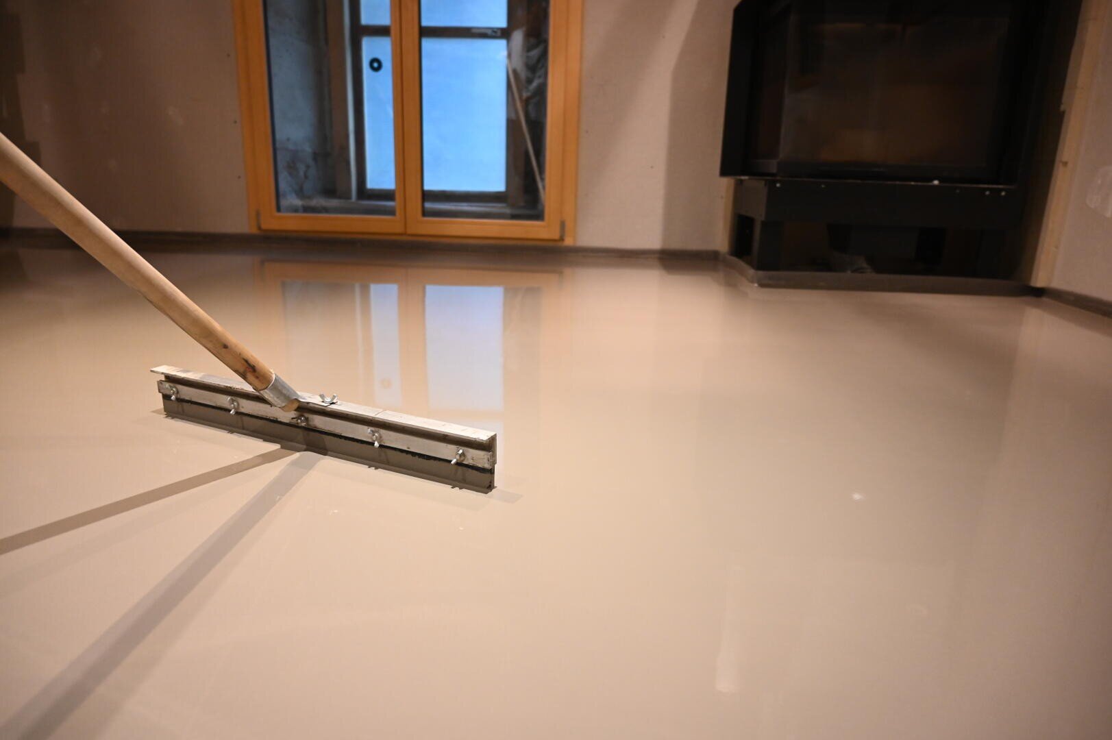 Nahaufnahme eines Fußbodens, der mit Fließestrich nivelliert und mit einem Werkzeug mit langem Stiel in einem Raum mit Kamin und großem Fenster geglättet wird.