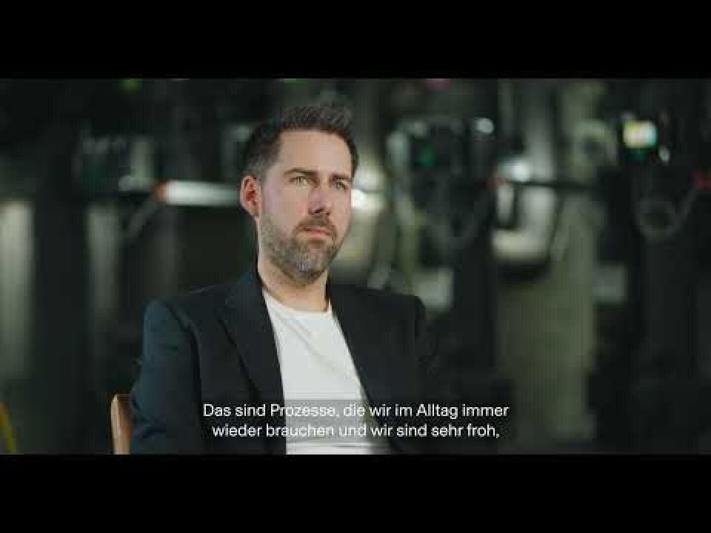 Ein Mann mit kurzem Haar und gestutztem Bart, der einen dunklen Blazer über einem weißen Hemd trägt, sitzt in einem Haus. Am unteren Rand des Bildes erscheinen deutsche Untertitel. Der Hintergrund ist unscharf und industriell.