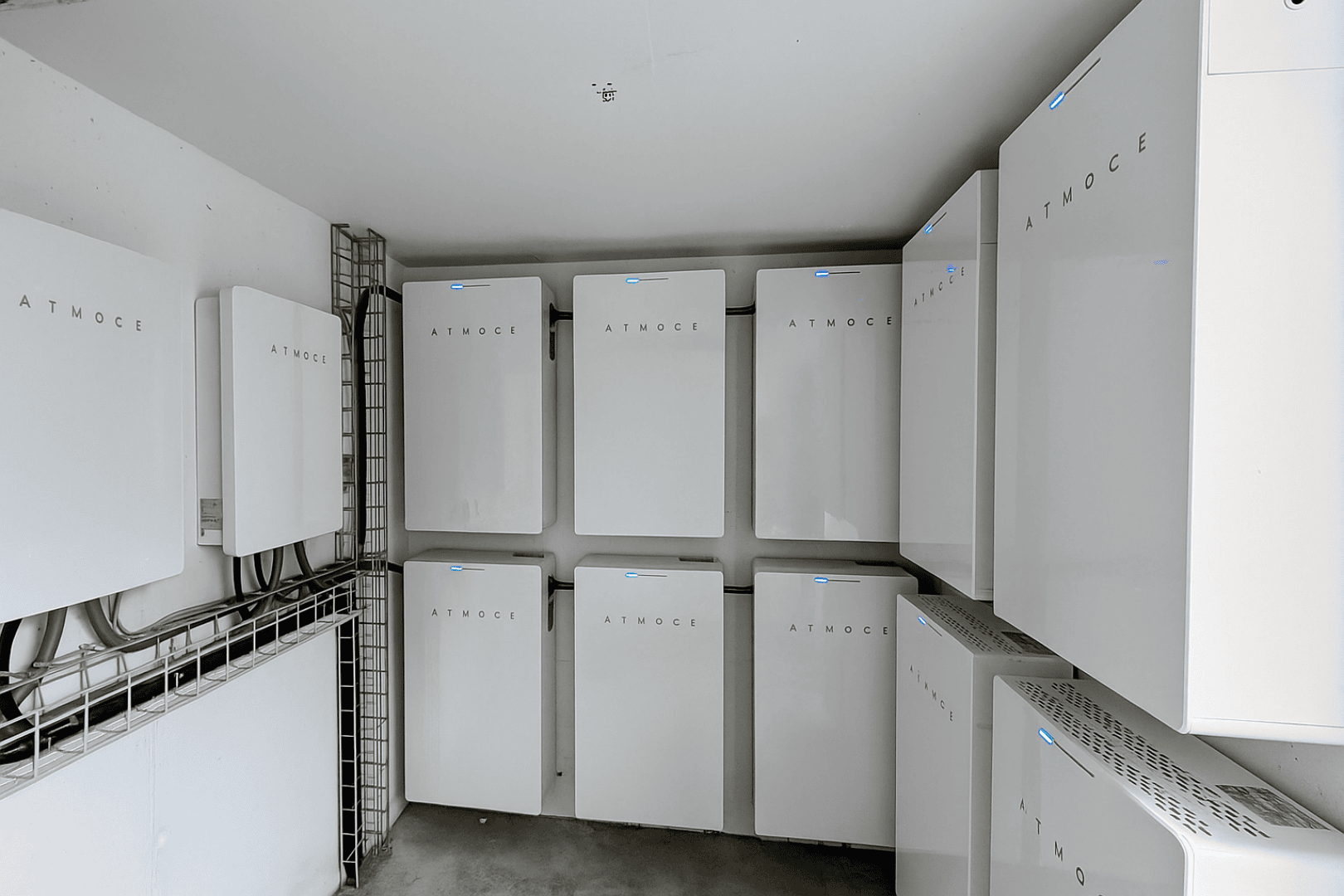 In einem sauberen, aufgeräumten Raum sind mehrere weiße, rechteckige Batteriespeicher mit der Aufschrift ATMOCE an den Wänden angebracht, von denen einige mit kleinen blauen Kontrollleuchten ausgestattet sind.