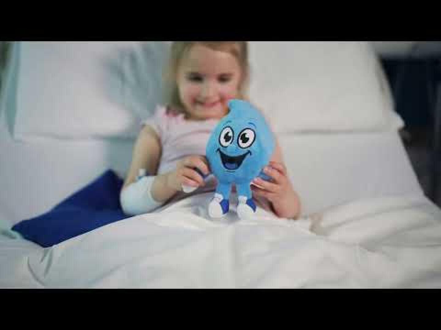 Ein junges Mädchen liegt in einem Krankenhausbett, lächelt und hält ein blaues Plüschtier mit einem fröhlichen Gesicht in der Hand. Sie hat einen Verband am Arm und ist mit einer weißen Decke zugedeckt.