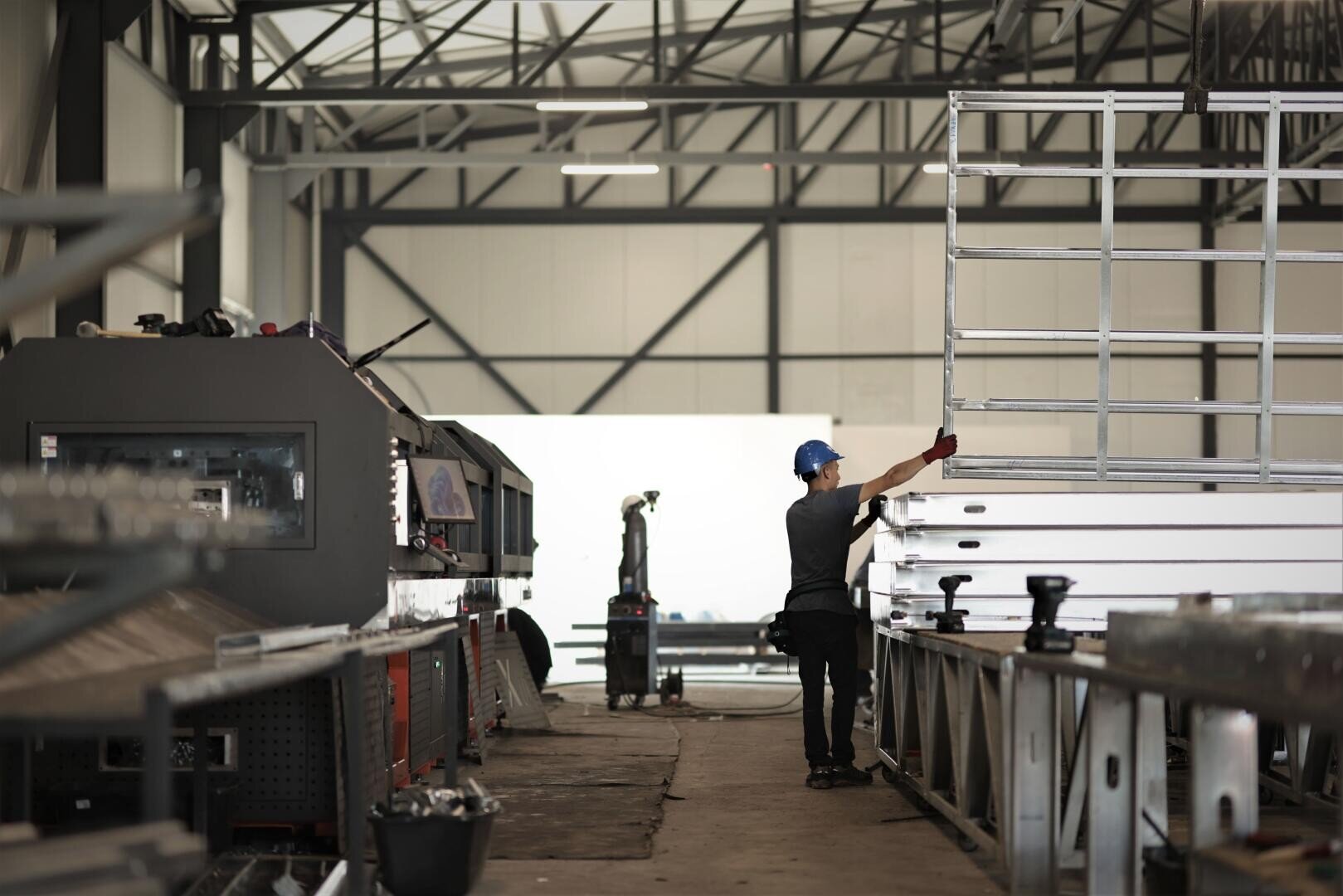 Ein Arbeiter mit einem blauen Sicherheitshelm und Handschuhen hantiert mit Metallrahmen im Inneren einer Industrieanlage, wobei im Hintergrund Maschinen und Stahlträger zu sehen sind.