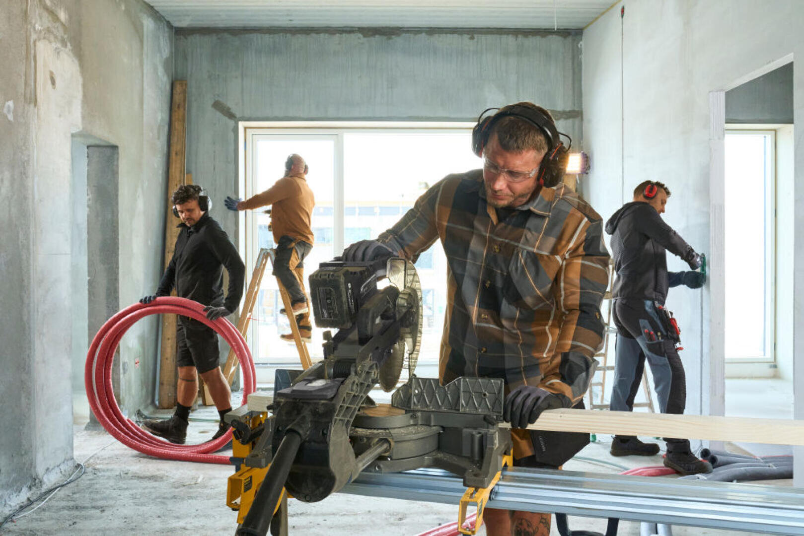 Quatre ouvriers du bâtiment sont en train de rénover une pièce. L'un travaille avec une scie à bois, un autre avec des tuyaux rouges, un troisième se tient sur une échelle et le quatrième travaille près d'une fenêtre. Tous portent des vêtements de protection. La pièce semble inachevée.