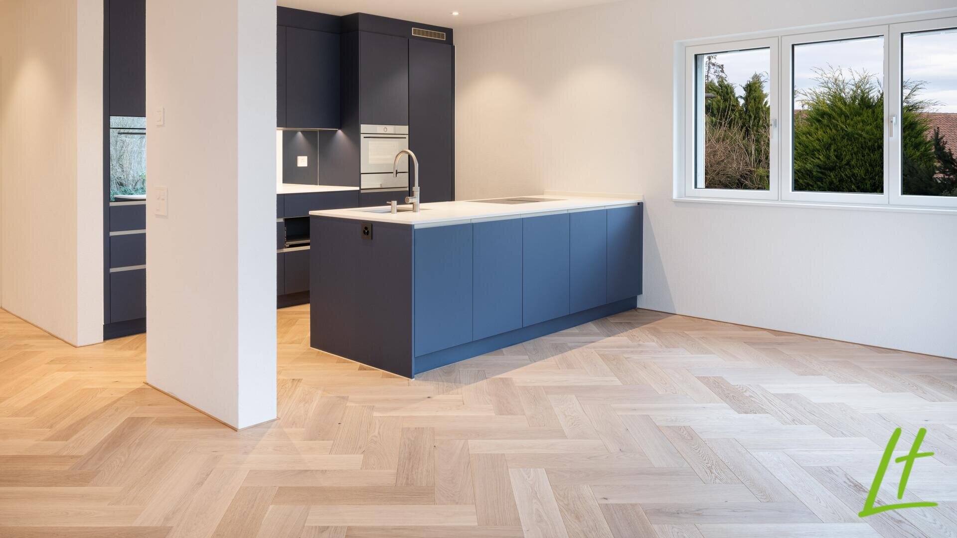 Moderne Küche mit blauen Schränken, weißen Arbeitsplatten, Einbaugeräten und hellem Holzfußboden im Fischgrätmuster. Ein großes Fenster lässt natürliches Licht herein, und in der unteren rechten Ecke befindet sich ein grünes "lt"-Logo.