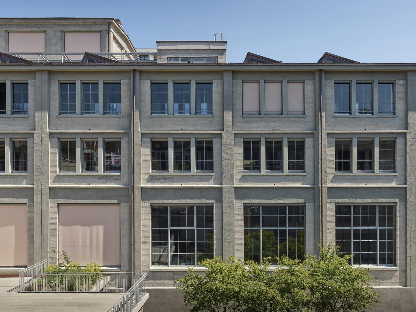 Ein modernes, mehrstöckiges Gebäude mit großen Fenstern und einer hellgrauen Fassade. Bei einigen Fenstern sind die rosa Jalousien teilweise zugezogen. Im Vordergrund sind Bäume und eine kleine Terrasse unter einem klaren blauen Himmel zu sehen.