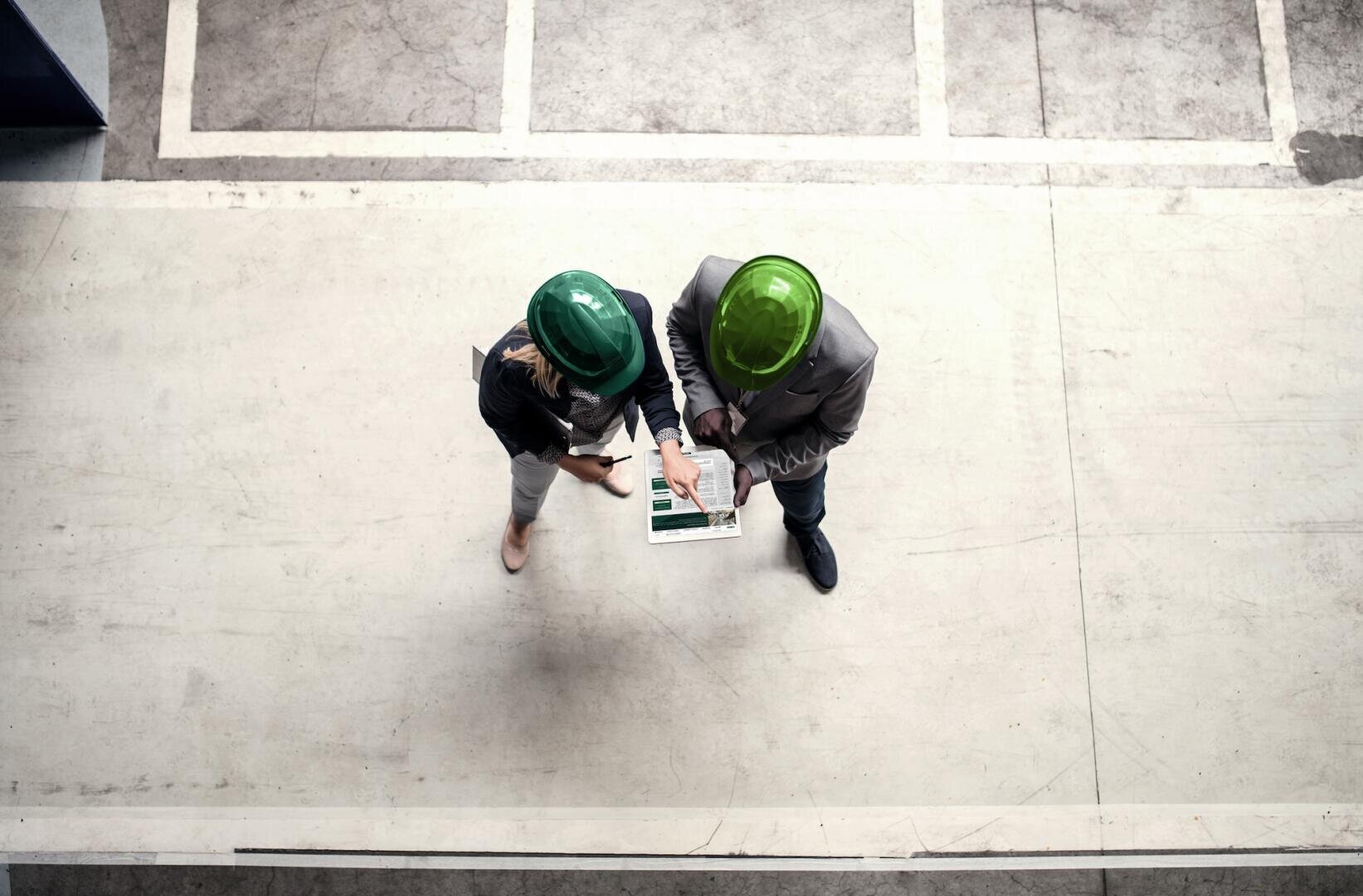 Zwei Personen mit grünen Schutzhelmen prüfen Dokumente oder Pläne auf einem Klemmbrett, während sie auf einem Betonboden stehen, von oben gesehen in einer Industrie- oder Bauumgebung.