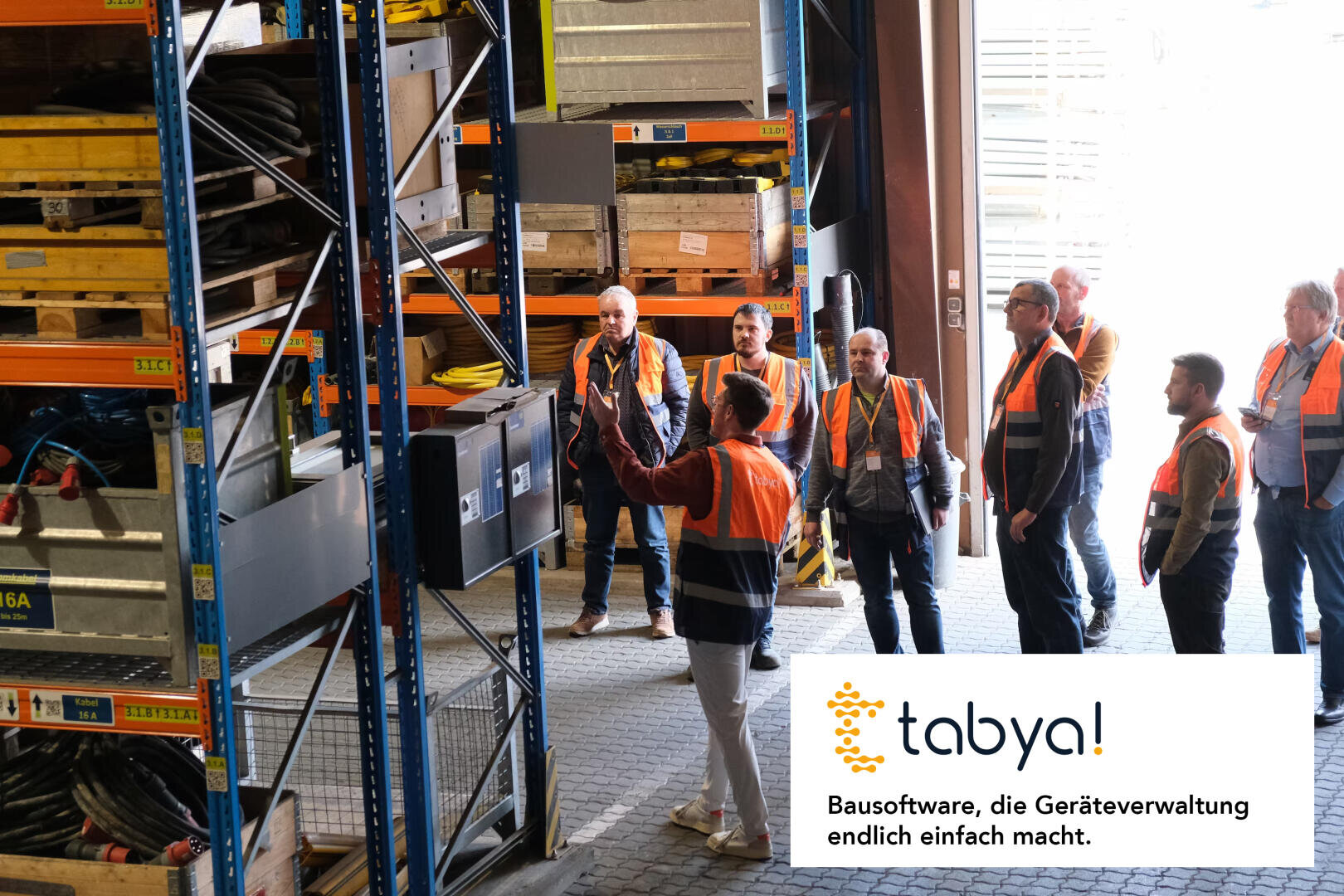 Eine Gruppe von Arbeitern in orangefarbenen Sicherheitswesten hört einem Vortragenden in einem Lagerhaus zu. Regale mit Geräten und Werkzeugen sind zu sehen. Das tabya!-Logo und der deutsche Text sind in der unteren rechten Ecke zu sehen.