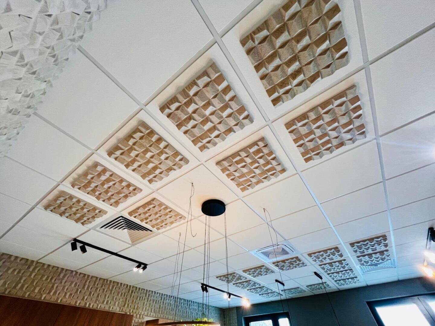 Eine Decke mit quadratischen Akustikplatten, die in einem Rastermuster angeordnet sind und strukturierte Oberflächen aufweisen. Auch Beleuchtungskörper und Lüftungsöffnungen sind sichtbar, wobei das natürliche Licht aus den darunter liegenden Fenstern kommt.