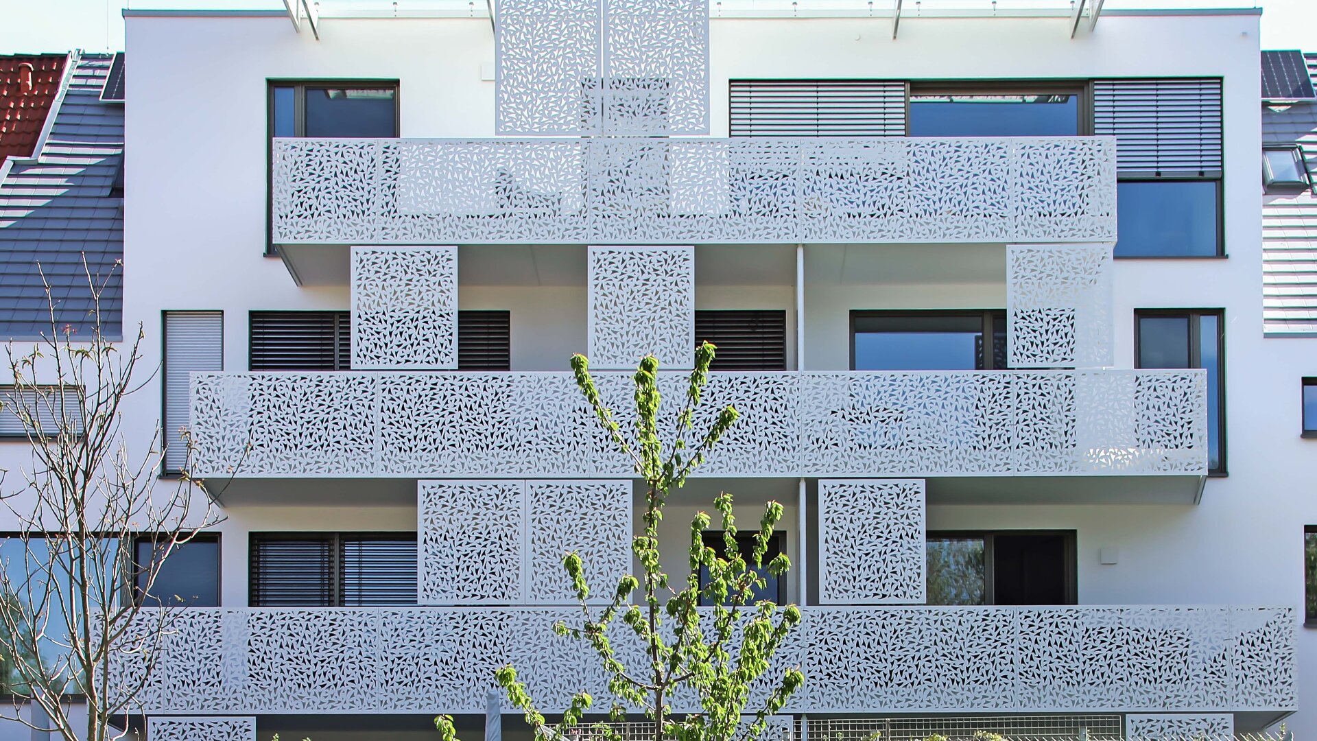 Modernes Wohnhaus mit weißem Balkongeländer aus perforiertem Metall und großen Fenstern. Ein kleiner Baum mit grünen Blättern steht im Vordergrund. Das Gebäude hat ein klares, minimalistisches Design.