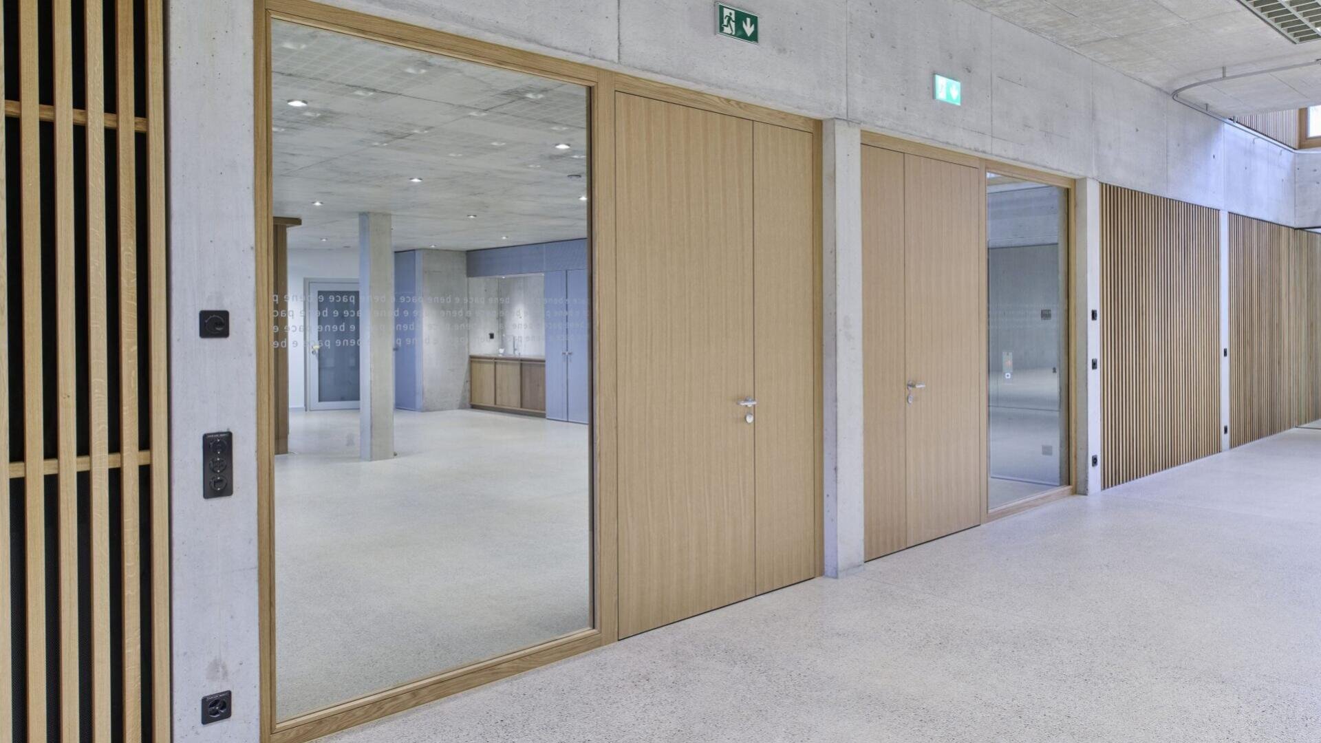 Ein moderner, minimalistischer Flur mit hellen Holztüren und großen Glasflächen. Die Böden und Wände sind in hellen Farben gehalten, und an der Decke befindet sich Sichtbeton. Ein leerer Raum mit Säulen ist durch das Glas sichtbar.