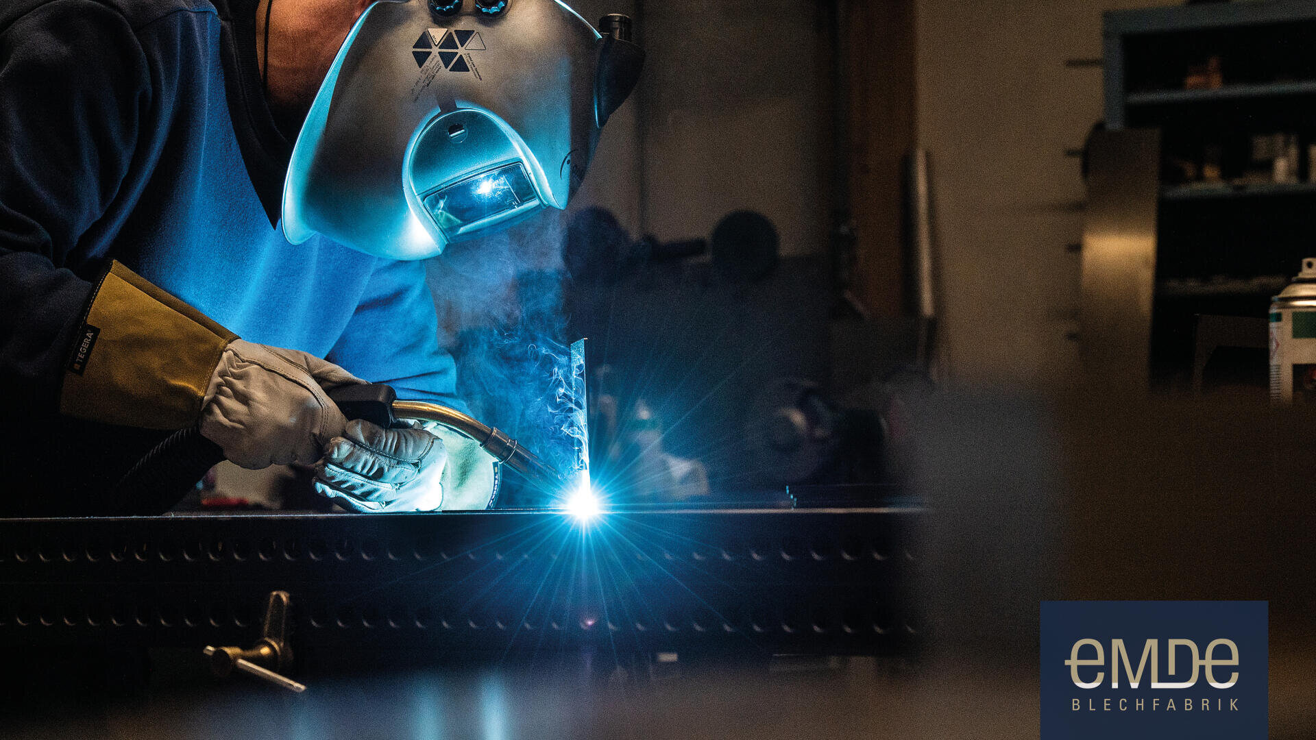 Eine Person, die einen Schweißerhelm und Handschuhe trägt, schweißt Metall und erzeugt dabei helle Funken. Im Hintergrund ist eine Werkstatt zu sehen, und in der unteren rechten Ecke erscheint das Logo der "eMDe Blechfabrik".