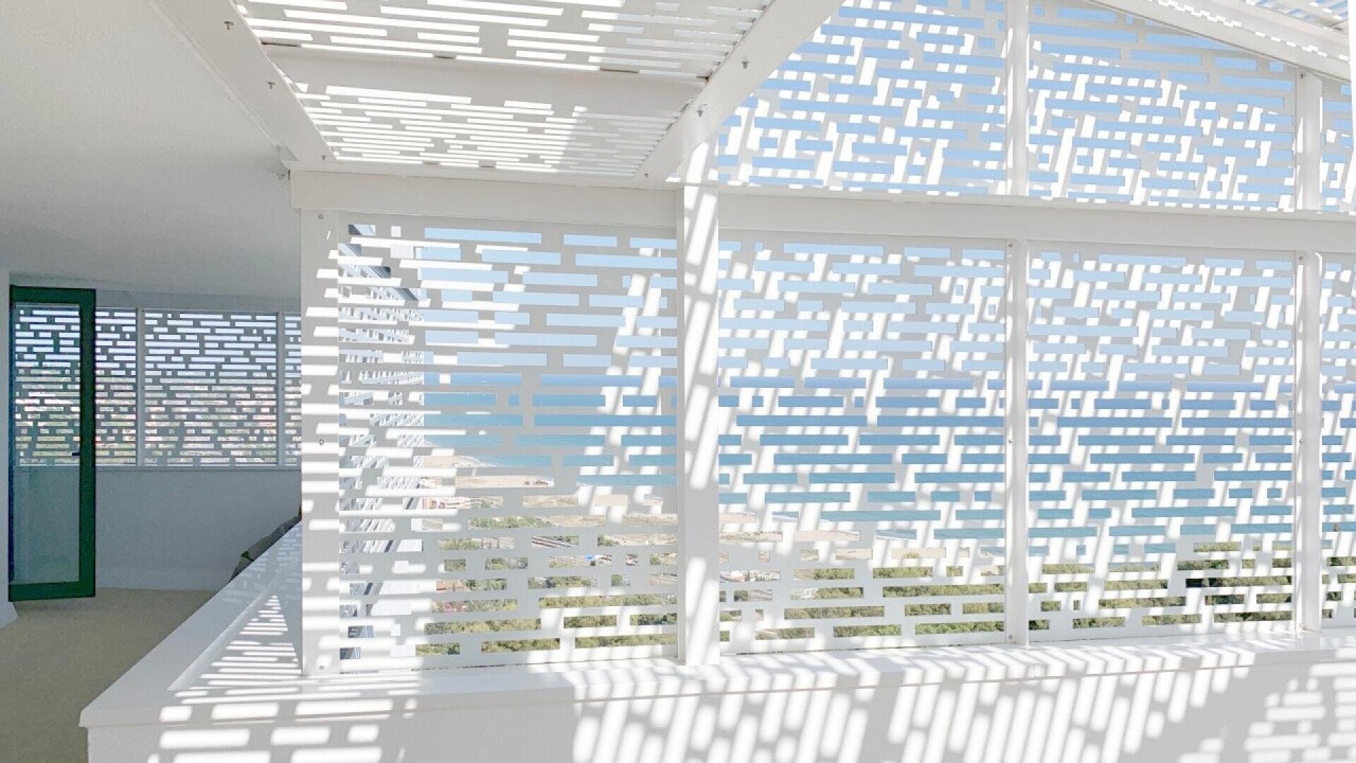 Un balcon lumineux présente des panneaux perforés blancs modernes qui projettent des ombres géométriques, avec une vue sur l'océan bleu et le littoral visible à travers les ouvertures. La lumière du soleil crée un motif sur le sol et les murs.