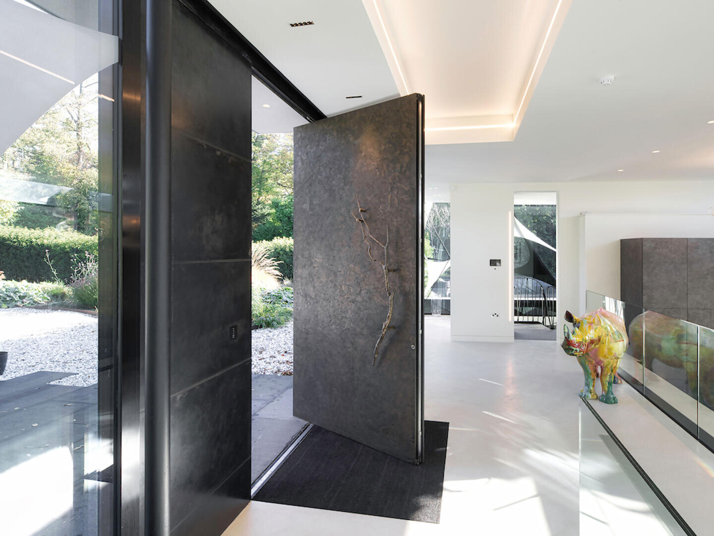 Entrée de maison moderne avec une grande porte battante sombre partiellement ouverte sur un jardin aménagé. L'intérieur se caractérise par des murs blancs sobres, une décoration minimale et une sculpture d'animal colorée sur le côté droit.