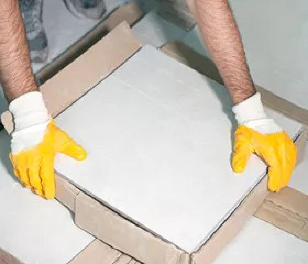 Eine Person, die gelbe Gummihandschuhe trägt, hebt oder legt eine große weiße Fliese in einen Karton.