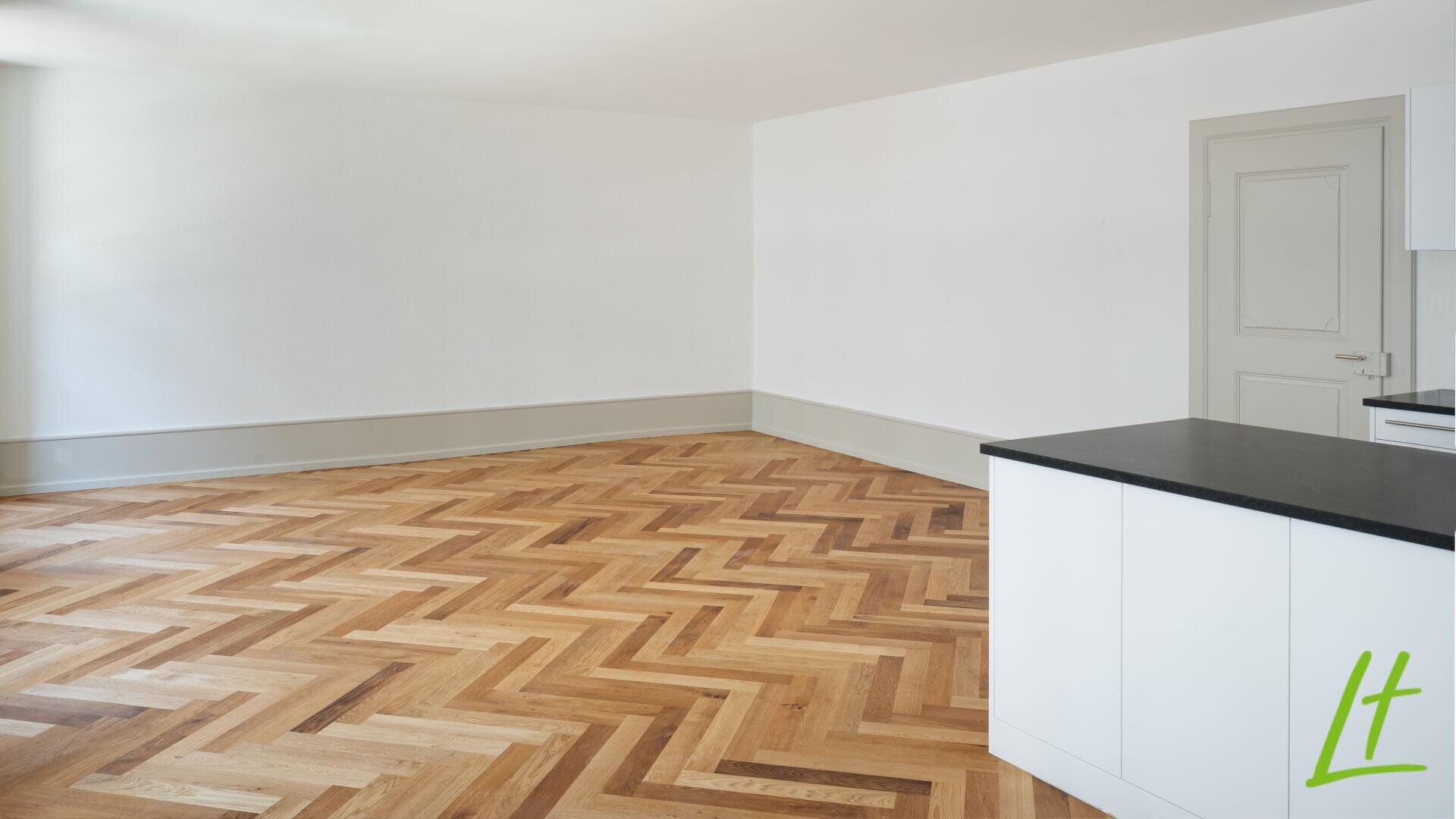 Ein heller, leerer Raum mit weißen Wänden, einem Holzboden mit Fischgrätenmuster, einer weißen Tür und einer weißen Küchenzeile mit einer schwarzen Arbeitsplatte im Vordergrund.